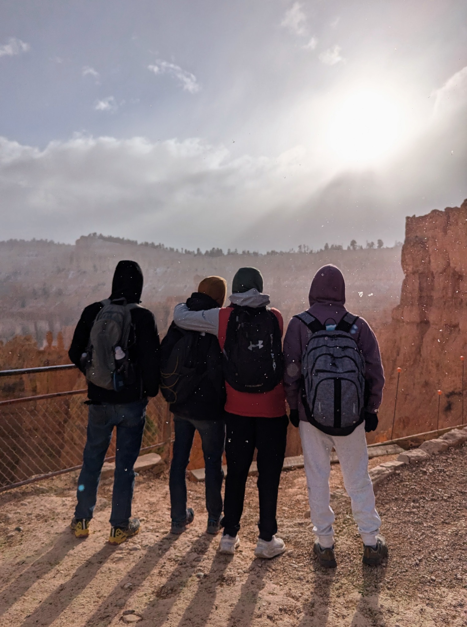 Four friends stand together beneath falling snow and sunlight during a birthday trip through Utah.