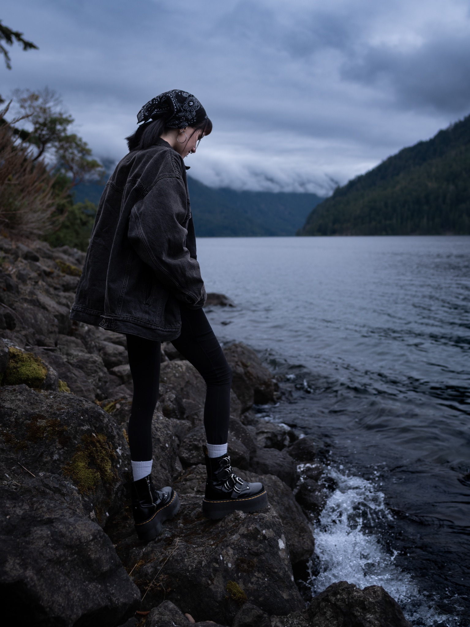 A woman steps along dark lakeshore rocks as water splashes beneath her boots.