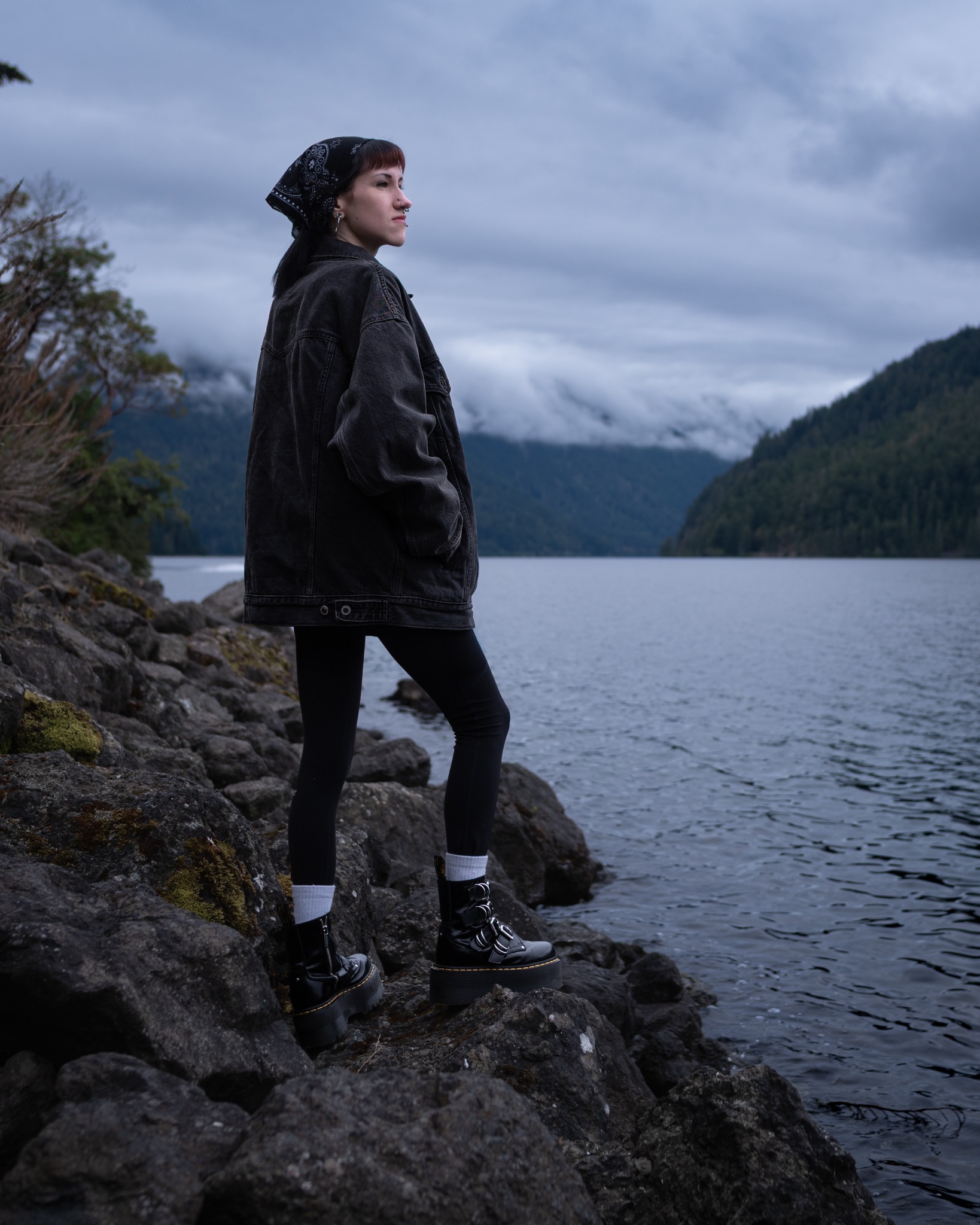 A woman stands in profile above a cold mountain lake under low clouds.